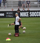 A person in sports attire stands on a soccer field, surrounded by several soccer balls and a carrier with bottles. The field is bordered by advertisements for Nike Soccer and app stores. Several people sit in bleachers in the background.