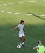 Athletes are on a soccer field engaging in practice. A player wearing a jersey with the number 14 is standing with a soccer ball at their feet, pointing to the right. Teammates are scattered around the field, some stretching or preparing for drills. A coach or staff member is carrying equipment, possibly looking towards the players.