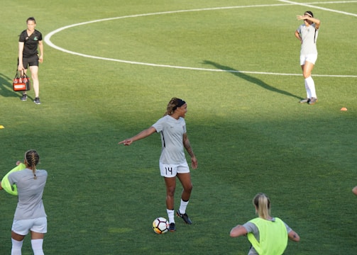Athletes are on a soccer field engaging in practice. A player wearing a jersey with the number 14 is standing with a soccer ball at their feet, pointing to the right. Teammates are scattered around the field, some stretching or preparing for drills. A coach or staff member is carrying equipment, possibly looking towards the players.