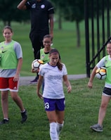A group of individuals on a grassy field, with some wearing sports attire including a white and purple jersey. One person is holding a soccer ball, and others are wearing bright green vests with shorts. A man in the background has a whistle, suggesting a coaching role.