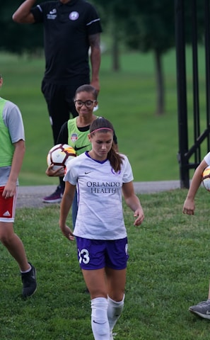 A group of individuals on a grassy field, with some wearing sports attire including a white and purple jersey. One person is holding a soccer ball, and others are wearing bright green vests with shorts. A man in the background has a whistle, suggesting a coaching role.