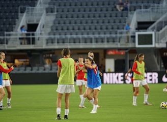 A group of female soccer players are on a field, wearing training bibs over their uniforms. They are practicing with a soccer ball, engaged in a passing drill. The stadium appears empty except for a few spectators and staff. The players are focused and in motion, showing dynamic movement.