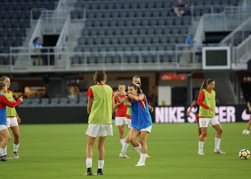 A group of female soccer players are on a field, wearing training bibs over their uniforms. They are practicing with a soccer ball, engaged in a passing drill. The stadium appears empty except for a few spectators and staff. The players are focused and in motion, showing dynamic movement.