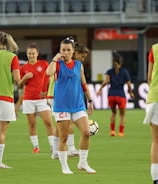 Several female soccer players are on a field during a practice session. They are wearing sports gear, including shirts, shorts, and training vests. Some players are conversing while one is holding a soccer ball. The players appear focused and ready, as they prepare for their practice or match.