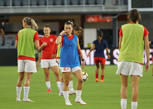Several female soccer players are on a field during a practice session. They are wearing sports gear, including shirts, shorts, and training vests. Some players are conversing while one is holding a soccer ball. The players appear focused and ready, as they prepare for their practice or match.