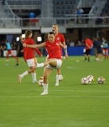 A group of soccer players in red jerseys and white shorts are practicing on a green field. One player in the foreground is stretching their leg, while others are engaged in warming up or training exercises. There are soccer balls scattered on the ground, and the setting appears to be a large sports stadium with empty stands in the background.