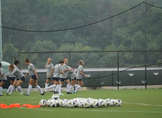 A group of female soccer players are engaged in a warm-up or training session on a grassy field. They are wearing coordinated gray shirts and shorts, with long white socks and soccer cleats. In the foreground, several soccer balls are neatly arranged on the ground. Behind them, a fence and a backdrop of lush green trees are visible, with netting also present.