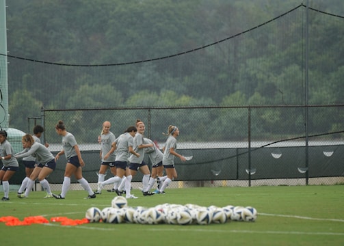 A group of female soccer players are engaged in a warm-up or training session on a grassy field. They are wearing coordinated gray shirts and shorts, with long white socks and soccer cleats. In the foreground, several soccer balls are neatly arranged on the ground. Behind them, a fence and a backdrop of lush green trees are visible, with netting also present.