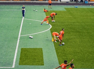 A group of people wearing bright orange sportswear are practicing on a green field. They appear to be athletes, likely soccer players, stretching and preparing for training. There are several soccer balls on the field, and a coach is observing from a distance. The field is surrounded by bleachers with a few onlookers seated, and the area is bordered by trees and residential buildings in the background.