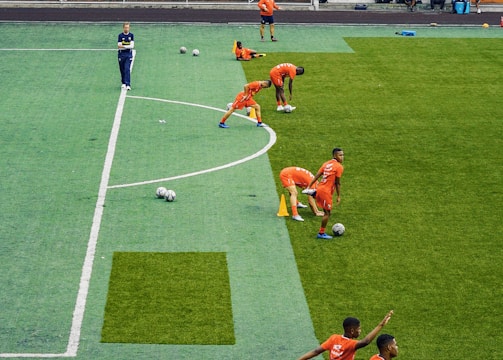 A group of people wearing bright orange sportswear are practicing on a green field. They appear to be athletes, likely soccer players, stretching and preparing for training. There are several soccer balls on the field, and a coach is observing from a distance. The field is surrounded by bleachers with a few onlookers seated, and the area is bordered by trees and residential buildings in the background.