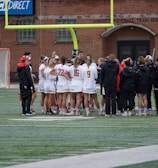 A group of athletes wearing sports uniforms huddles on a grassy field, likely preparing or strategizing for a game. The players are surrounded by additional team members or coaches wearing black jackets. A goalpost and net are visible in the background, along with a brick building.