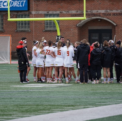 A group of athletes wearing sports uniforms huddles on a grassy field, likely preparing or strategizing for a game. The players are surrounded by additional team members or coaches wearing black jackets. A goalpost and net are visible in the background, along with a brick building.