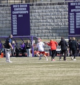 A sports team is practicing or playing on a grassy field in front of a large stone wall with banners displaying men's soccer achievements. People are wearing sports attire, and some are engaged in conversation or coaching.