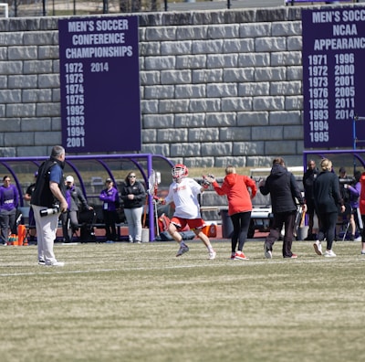 A sports team is practicing or playing on a grassy field in front of a large stone wall with banners displaying men's soccer achievements. People are wearing sports attire, and some are engaged in conversation or coaching.