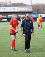 A group of people are on a soccer field, with some wearing sports attire. In the foreground, a person in red sports clothing is walking alongside someone in a dark tracksuit. Other people are visible in the background, some wearing yellow jerseys. A building with advertising banners, including a prominent sign reading 'Ready, Set, Smile,' is in the background.