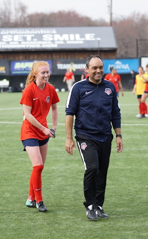 A group of people are on a soccer field, with some wearing sports attire. In the foreground, a person in red sports clothing is walking alongside someone in a dark tracksuit. Other people are visible in the background, some wearing yellow jerseys. A building with advertising banners, including a prominent sign reading 'Ready, Set, Smile,' is in the background.