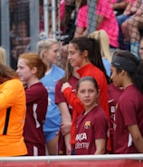 A group of young female athletes in sports attire are gathered together, some wearing red jackets or maroon jerseys with the logo of a soccer academy. They appear to be standing in front of a wire fence with spectators in the background, dressed in casual clothing.