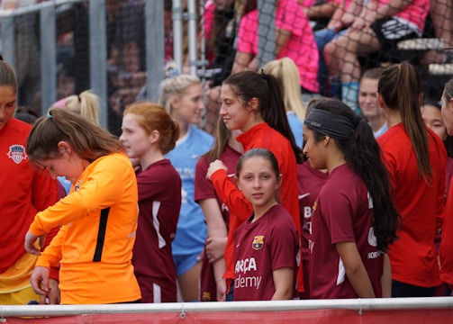 A group of young female athletes in sports attire are gathered together, some wearing red jackets or maroon jerseys with the logo of a soccer academy. They appear to be standing in front of a wire fence with spectators in the background, dressed in casual clothing.