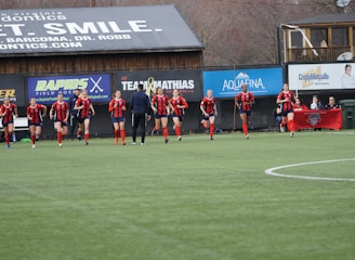 A group of soccer players in red and black uniforms are running on a green field towards the camera. A coach or trainer stands in front of them, possibly giving instructions. Advertising boards and a banner are visible in the background, along with some spectators seated at the sidelines.
