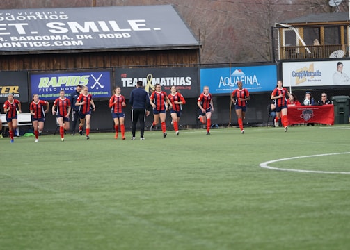 A group of soccer players in red and black uniforms are running on a green field towards the camera. A coach or trainer stands in front of them, possibly giving instructions. Advertising boards and a banner are visible in the background, along with some spectators seated at the sidelines.