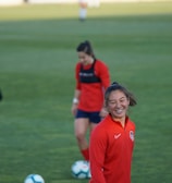 Players are on a grassy sports field, focusing on a woman in a red sports jersey smiling. Other individuals are present, wearing similar sports attire. Soccer balls are on the ground.