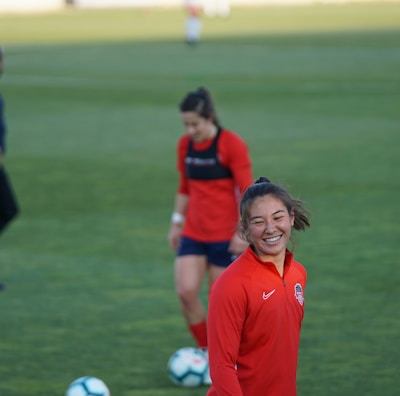 Players are on a grassy sports field, focusing on a woman in a red sports jersey smiling. Other individuals are present, wearing similar sports attire. Soccer balls are on the ground.