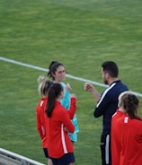A group of people wearing sports attire are gathered on a grassy field. Some individuals are dressed in red jackets, while others are in turquoise and navy blue. A coach appears to be giving instructions. Sports equipment, such as water bottles and a bench with towels, can be seen nearby.