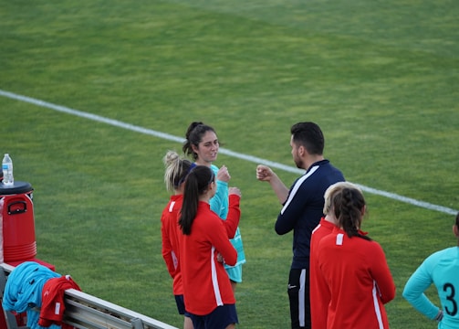 A group of people wearing sports attire are gathered on a grassy field. Some individuals are dressed in red jackets, while others are in turquoise and navy blue. A coach appears to be giving instructions. Sports equipment, such as water bottles and a bench with towels, can be seen nearby.