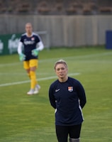A person stands on a soccer field, wearing a navy sports jacket with a logo. In the background, another person in sports gear, including yellow shorts, is visible, holding goalkeeper gloves. A third person is near some soccer balls, facing away. The setting appears to be a practice session with advertising boards visible along the sidelines.