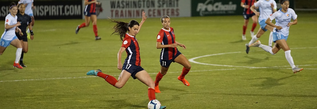 A group of female soccer players in action during a game on a grass field. One player wearing a red and navy uniform is about to kick the ball, while other players in similar uniforms and white uniforms are either running towards her or observing. The scene is set in an outdoor stadium with a sign indicating Washington Spirit Training Centers in the background.