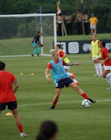 A group of female soccer players engaged in a practice session on a grass field. They wear red and blue uniforms, with some in training bibs. A visible goalpost, cones, and soccer balls are scattered around. In the background, a few spectators watch from sidelines near a banner with logos.