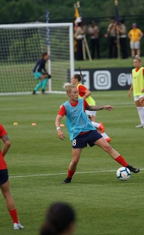 A group of female soccer players engaged in a practice session on a grass field. They wear red and blue uniforms, with some in training bibs. A visible goalpost, cones, and soccer balls are scattered around. In the background, a few spectators watch from sidelines near a banner with logos.