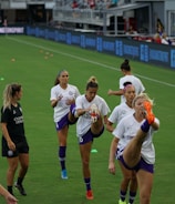 A group of female soccer players in matching uniforms is stretching on a soccer field. They are practicing high kicks as part of their warm-up routine. The grass is bright green, and there are small orange training cones scattered around. A coach or trainer in black attire is observing their practice.