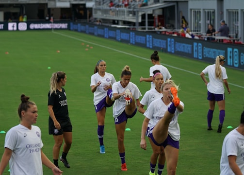 A group of female soccer players in matching uniforms is stretching on a soccer field. They are practicing high kicks as part of their warm-up routine. The grass is bright green, and there are small orange training cones scattered around. A coach or trainer in black attire is observing their practice.