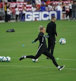 A soccer player in a black sports outfit is mid-action on a green field, focusing intently on kicking a soccer ball. Several soccer balls are scattered on the grass nearby, and a coach or teammate stands in the background watching. Spectators can be seen in the distant stands.