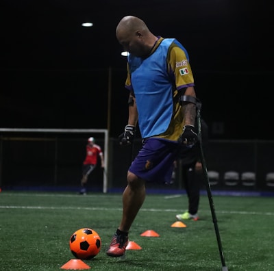 A man wearing a blue training vest is practicing on a football field. He is using crutches and moving a bright orange ball with his foot. Other people in sports attire can be seen in the background, and the field is marked with orange cones.