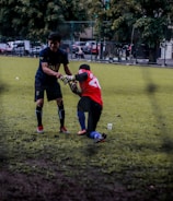 Two soccer players are on a grassy field; one is helping another off the ground. They are wearing different colored jerseys, indicating they might be from opposing teams or in training. Other players and some parked cars are visible in the background, suggesting an urban setting.