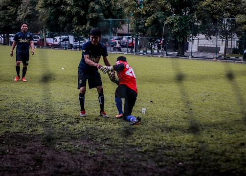 Two soccer players are on a grassy field; one is helping another off the ground. They are wearing different colored jerseys, indicating they might be from opposing teams or in training. Other players and some parked cars are visible in the background, suggesting an urban setting.