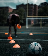 A person is engaged in a soccer training session on an outdoor field. The individual is focused on maneuvering through orange cones arranged on the grass. In the foreground, a soccer ball rests on the ground. There are buildings and trees in the background under a partly cloudy sky.