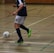 A person in a navy blue and white sports uniform is playing futsal in an indoor court. The individual is about to kick a colorful ball while wearing bright green and black shoes. The court is marked with various colored lines and has a wooden wall in the background.